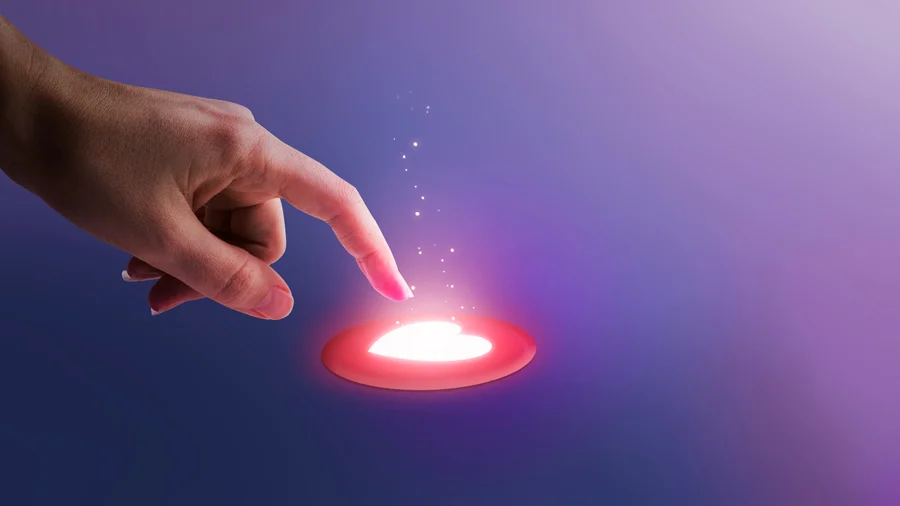 Mano que clica con un dedo un botón con forma de corazón iluminado sobre fondo lila. 